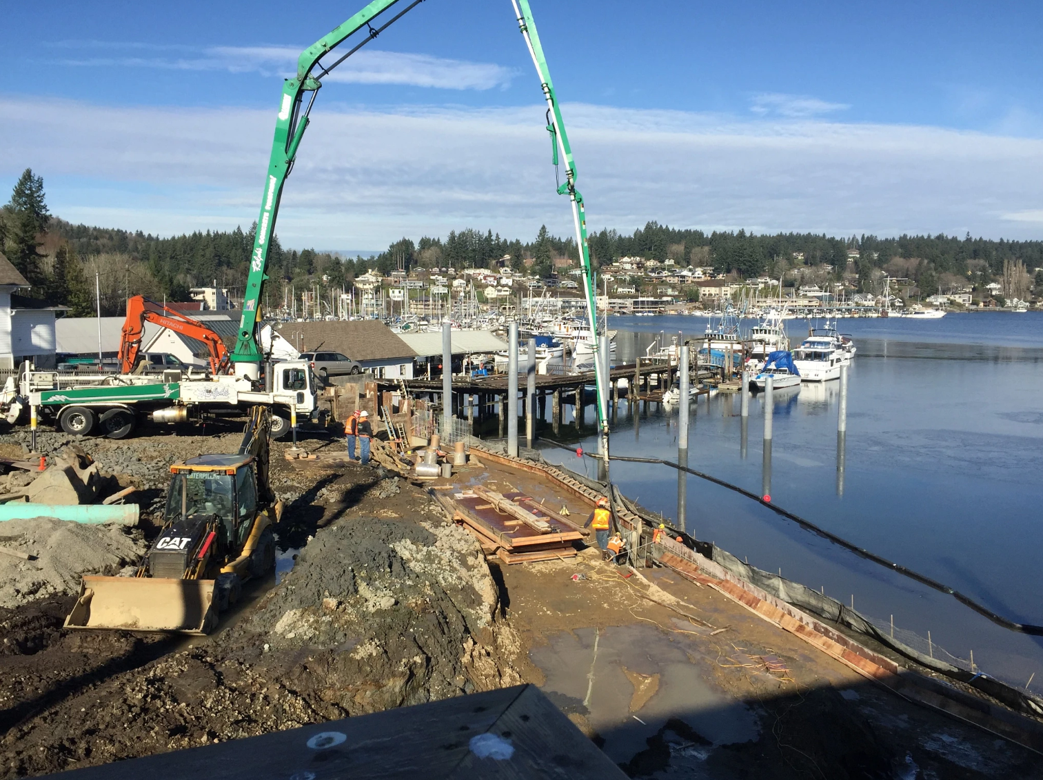 Construction work along a waterfront marina, with workers in safety gear operating heavy equipment, including a concrete pump truck and excavator, beside a muddy shoreline. Docks, pilings, and moored boats extend into calm water, with a small coastal town and tree-covered hills in the background under a clear sky.